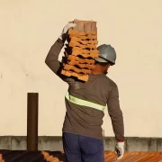 Operário da construção civil carregando tijolos nos ombros. Autora fala em normas de saúde do trabalhador.