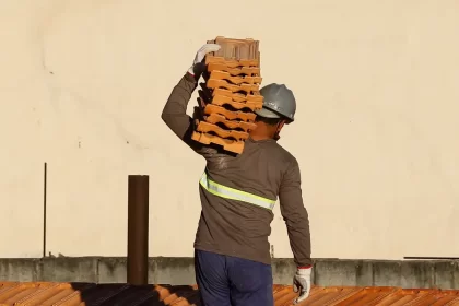 Operário da construção civil carregando tijolos nos ombros. Autora fala em normas de saúde do trabalhador.